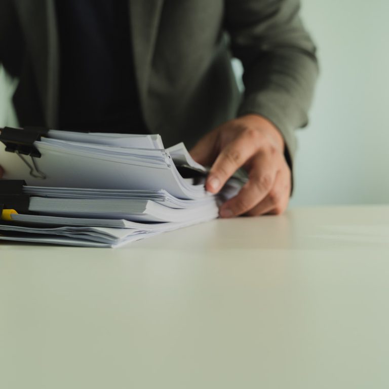 Stacks of paper files and checking document businessman checks financial documents on his laptop, managing corporate reports and legal paperwork at his office desk.